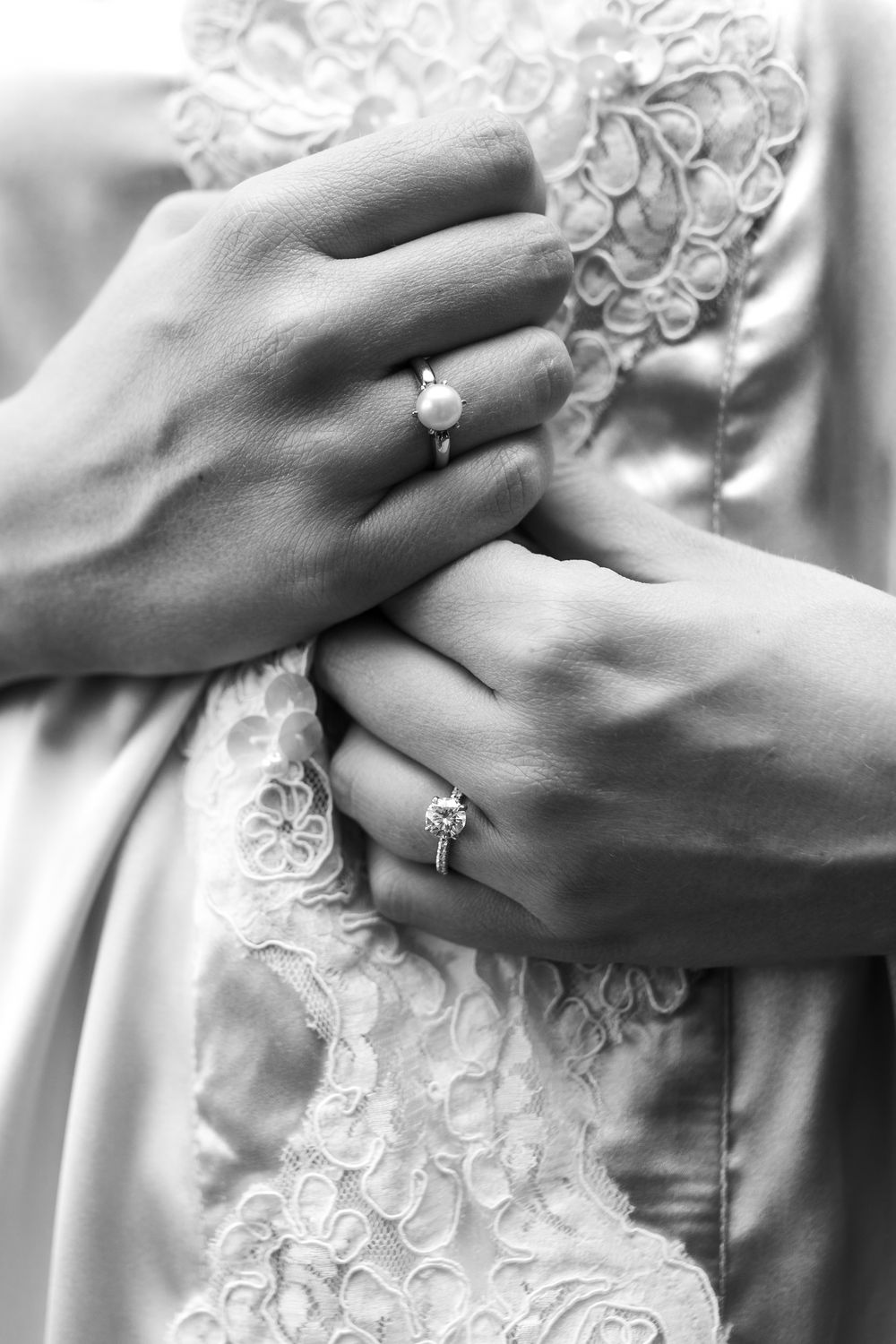 Black and white close-up photo of hands with wedding rings against delicate lace fabric details.