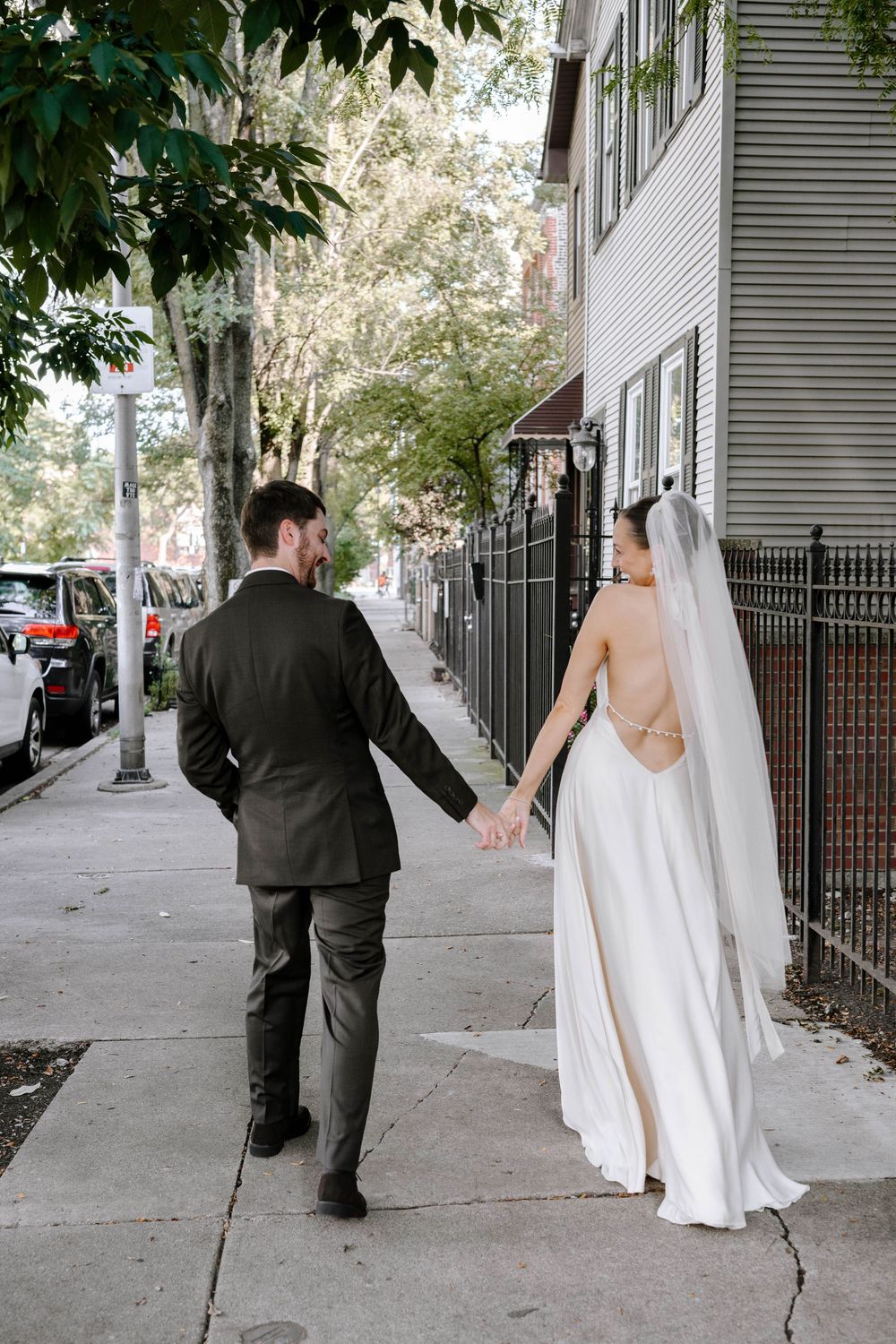 A couple in wedding attire playfully walks down a tree-lined urban sidewalk in sequence.