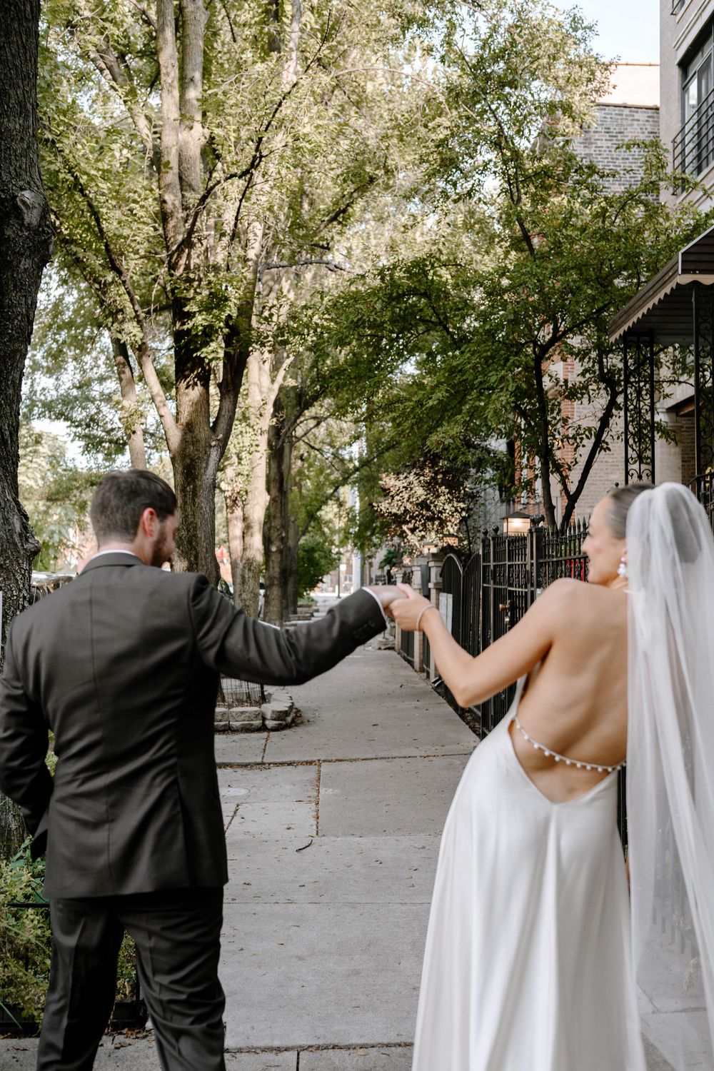 Newlywed couple walking down a tree-lined city sidewalk holding hands on their wedding day.
