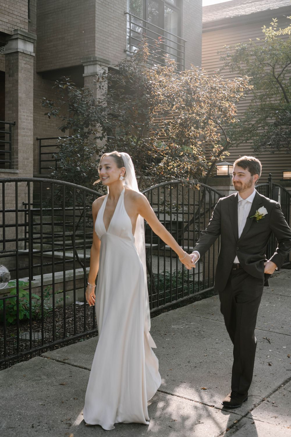 A couple in wedding attire holds hands while walking along a city sidewalk on a sunny day.