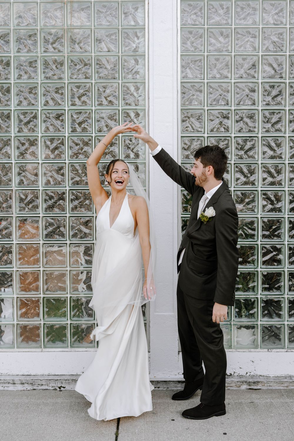 Newlyweds share a joyful dance pose against a glass block wall backdrop.