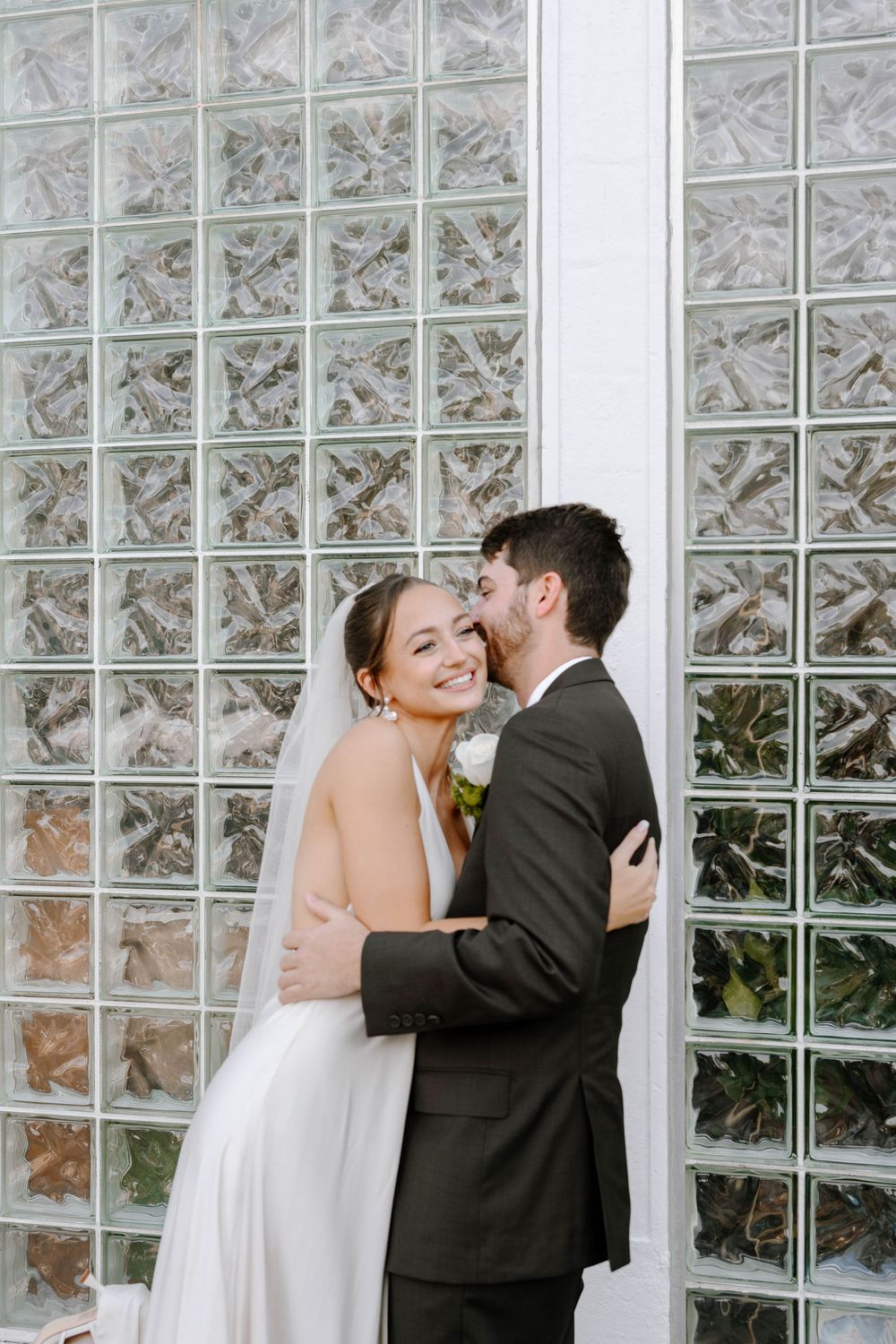 A romantic wedding couple shares a tender moment against a glass block wall backdrop.
