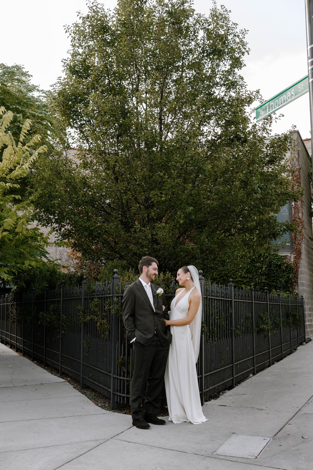 Elegant wedding couple poses on city corner with lush green trees and black iron fence in background.