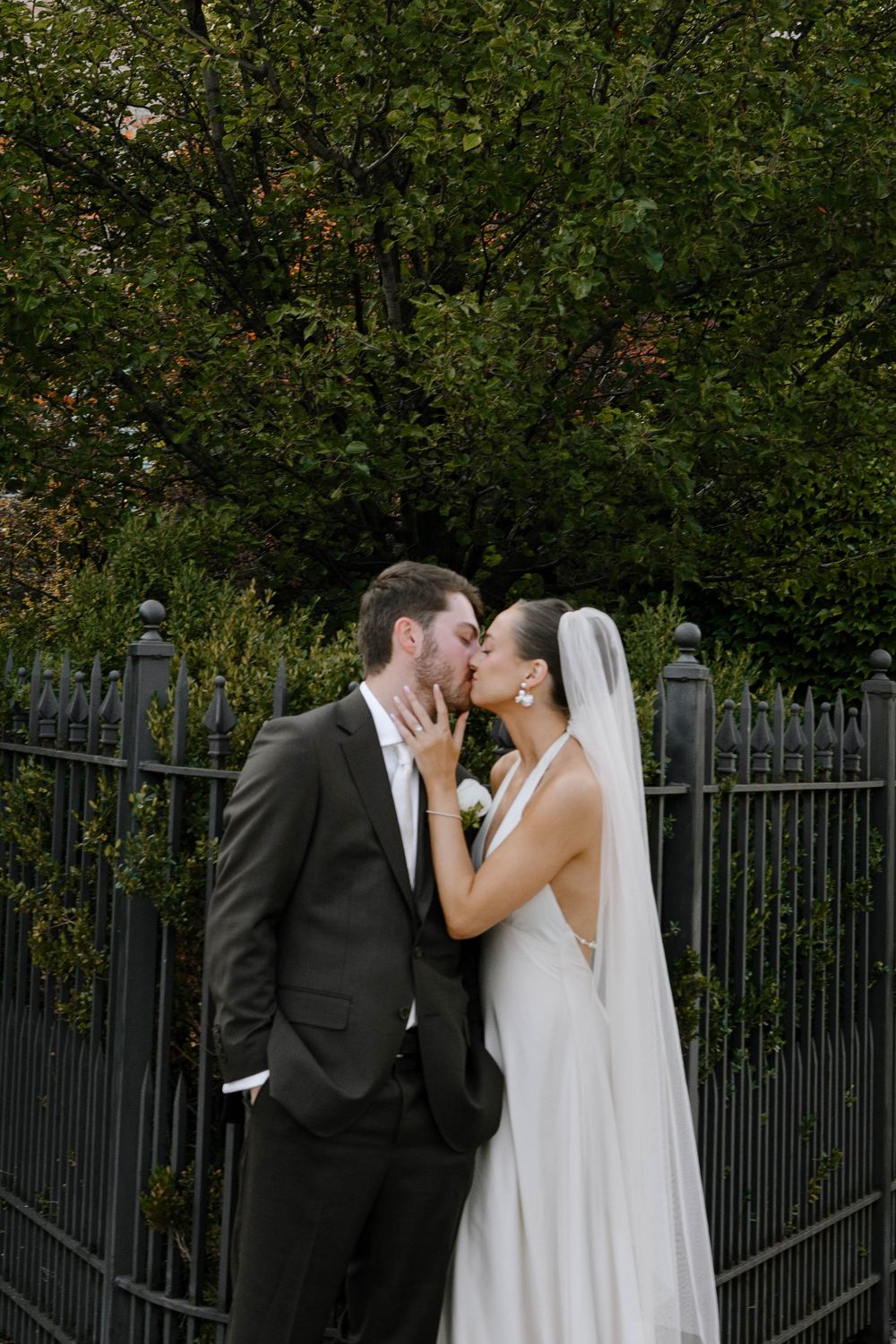 Romantic wedding couple embraces in front of an iron gate and lush garden.