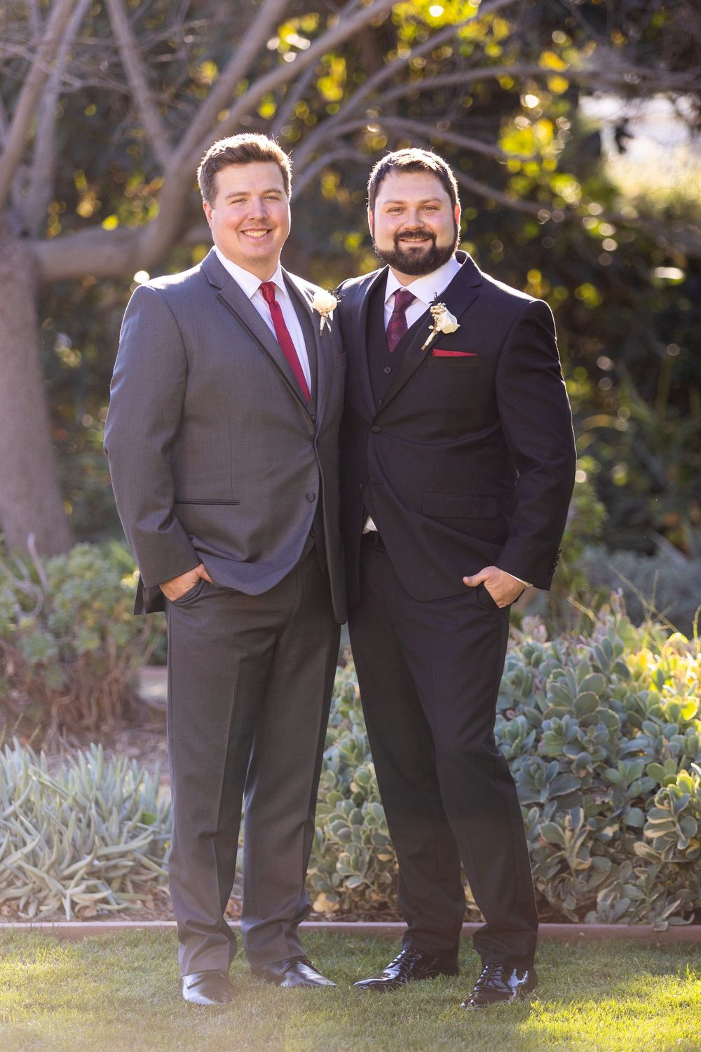 Two people in formal attire stand together outdoors with yellow flowers and greenery in the background.