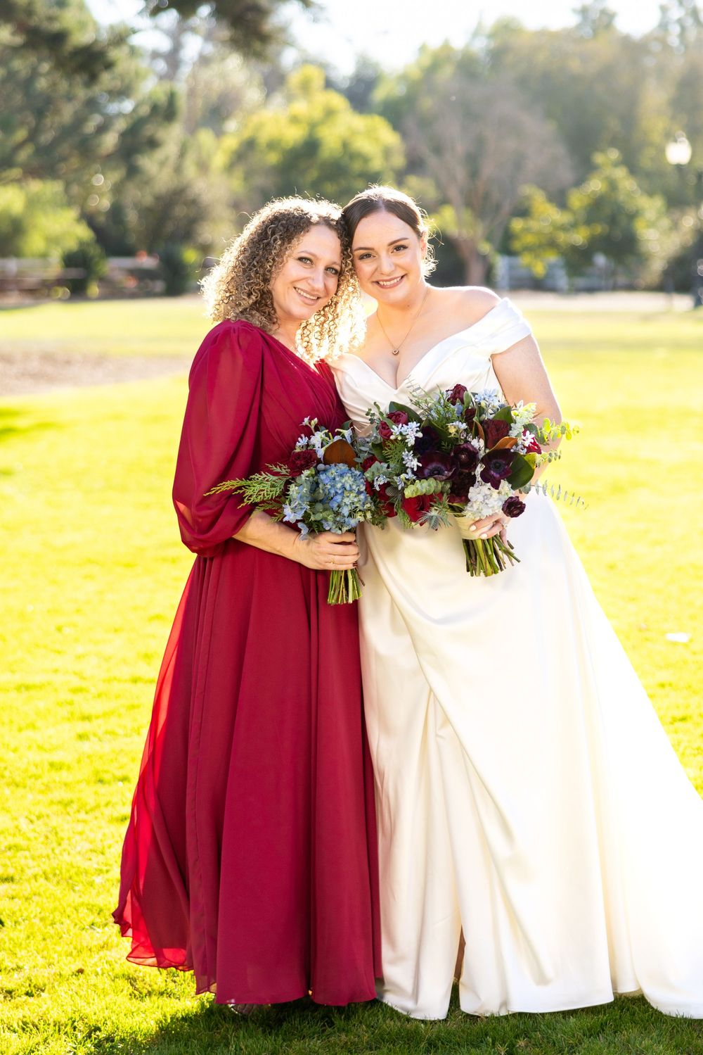 Two people in contrasting red and white dresses embrace while holding floral bouquets in a sunny outdoor field.