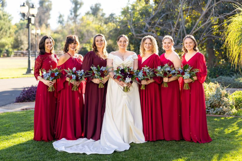 Bridesmaids in flowing red dresses hold vibrant bouquets next to bride in white gown outdoors.