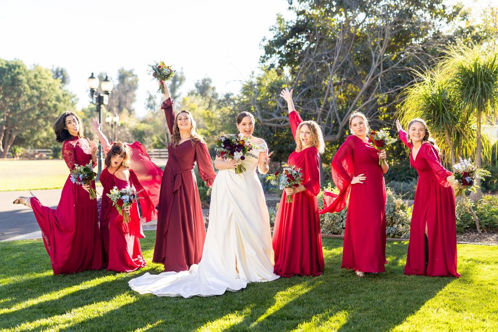 Wedding party in red dresses poses together on sunny lawn with palm trees in background.