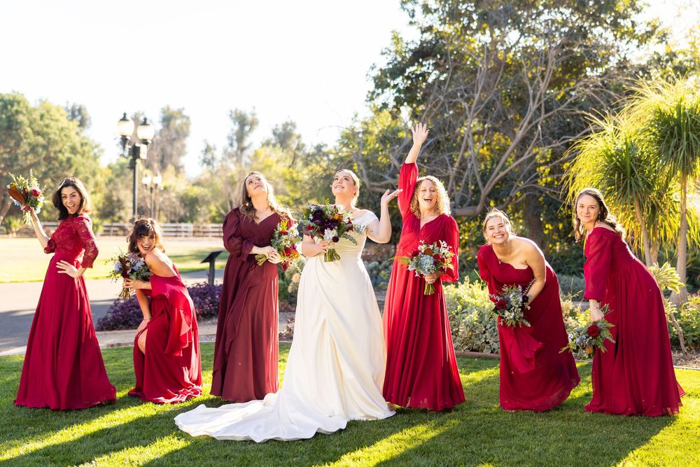 Wedding party in red bridesmaid dresses and white bridal gown pose together outdoors in a sunny garden setting.