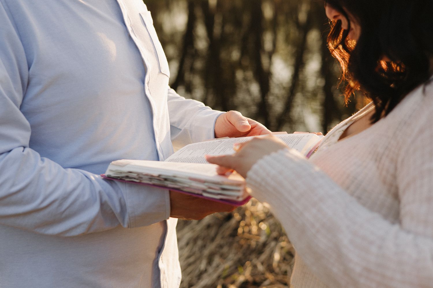 Close-up of couple holding hands while exchanging vows outdoors, with soft golden light filtering through trees.