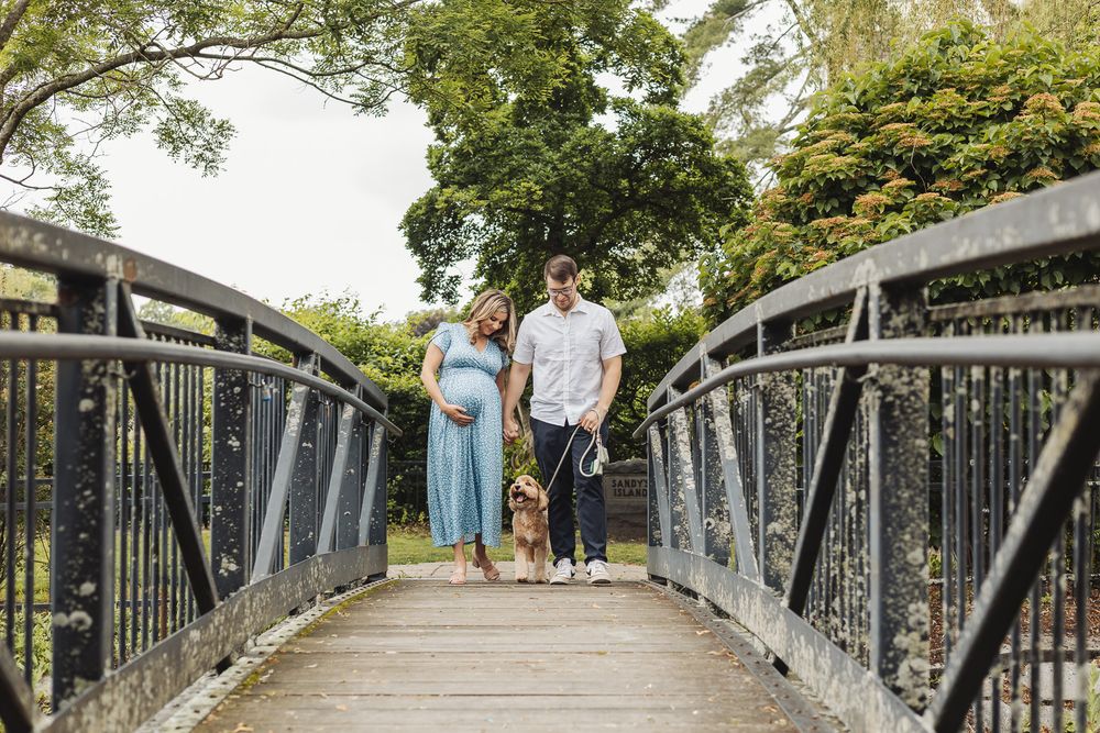 Couple walking along a scenic metal footbridge surrounded by lush green trees on a sunny day.