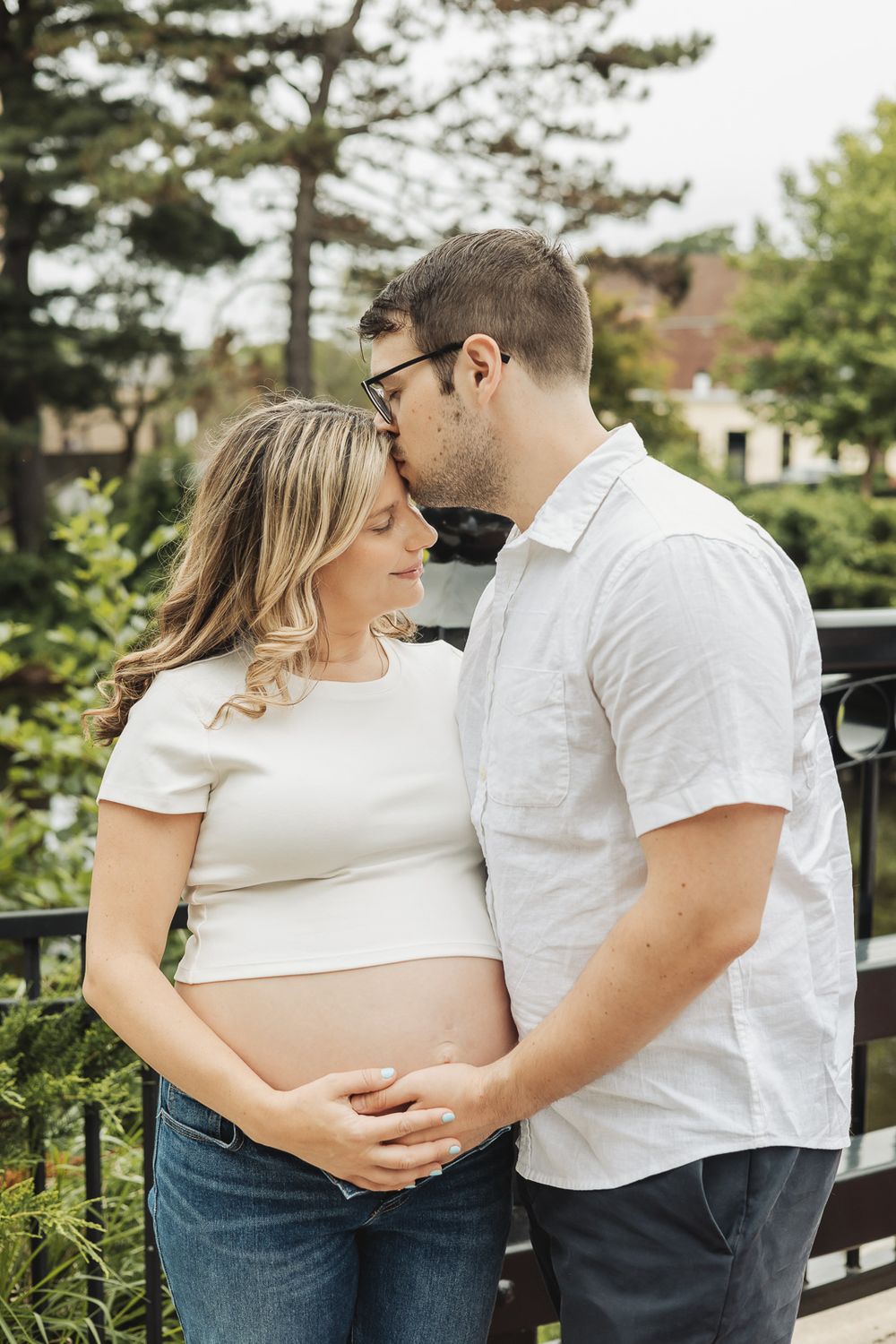 A couple in white shirts embraces tenderly during a maternity photoshoot in an outdoor setting.