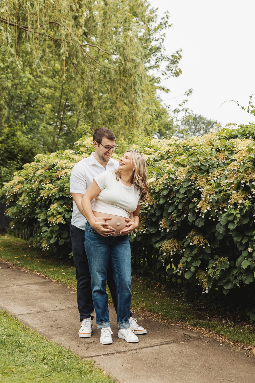 Expecting couple embraces on garden path surrounded by lush greenery and flowering bushes.