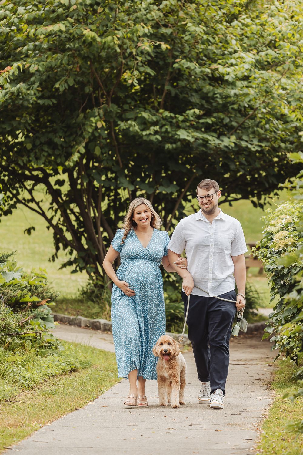 A couple walks their goldendoodle dog along a tree-lined path on a sunny summer day.