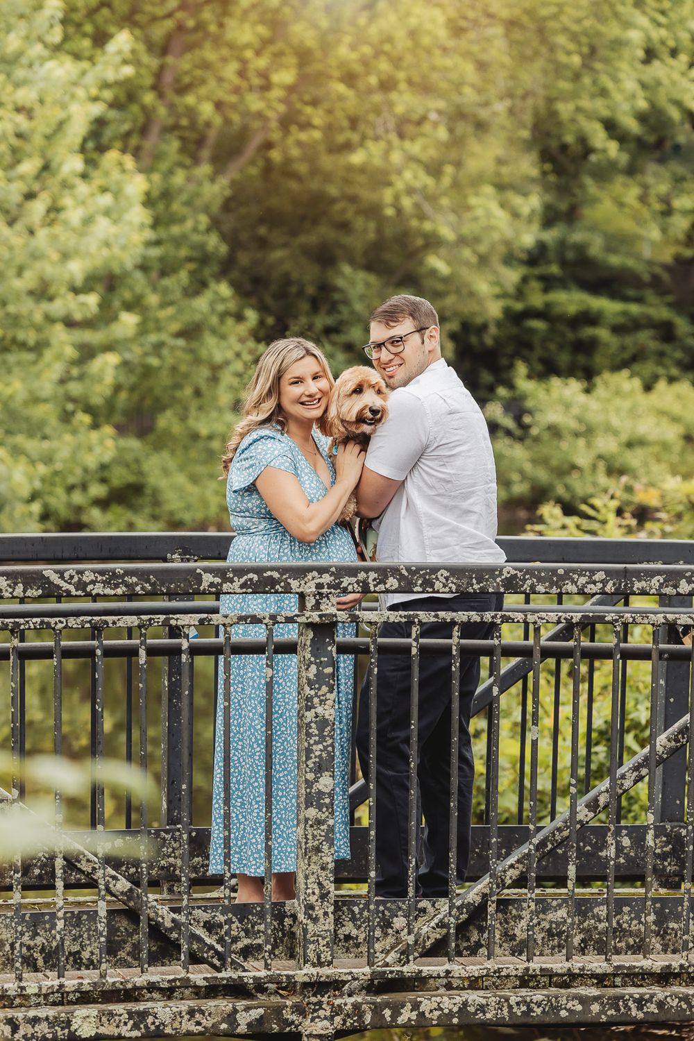 A couple shares a romantic moment together on a black iron bridge surrounded by lush green foliage.