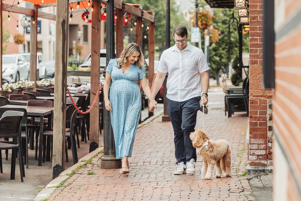 Couple walking their golden retriever on brick sidewalk near outdoor restaurant seating on sunny day.