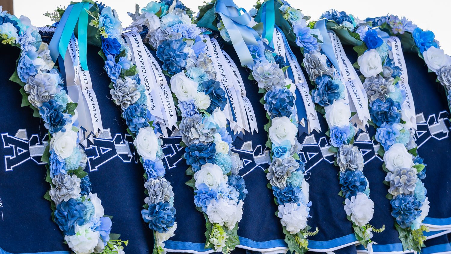 Elegant floral leis in shades of blue and white hang in a row against a dark blue background.