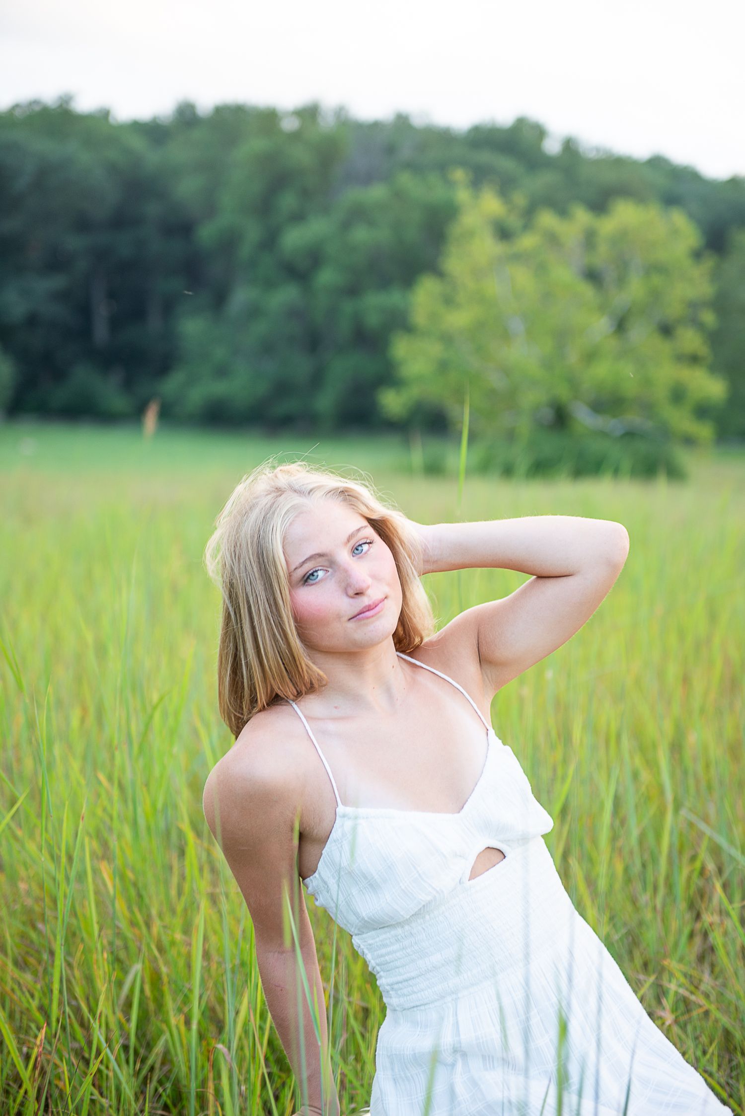 A woman in a white dress poses gracefully in a lush green field on a summer day.
