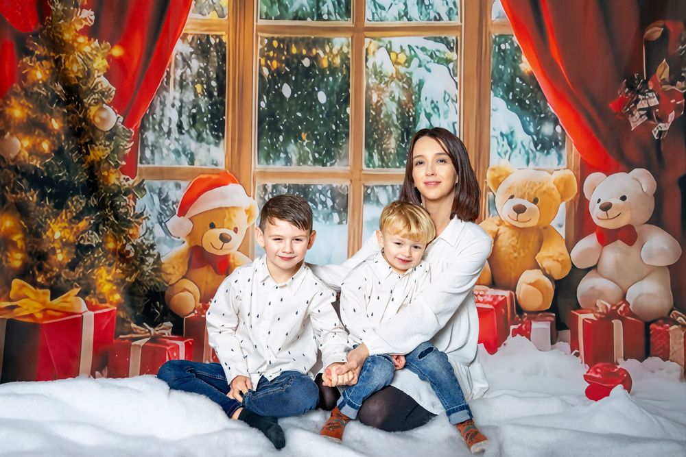 Children in white pajamas sit among holiday decorations and teddy bears near a window with red curtains.