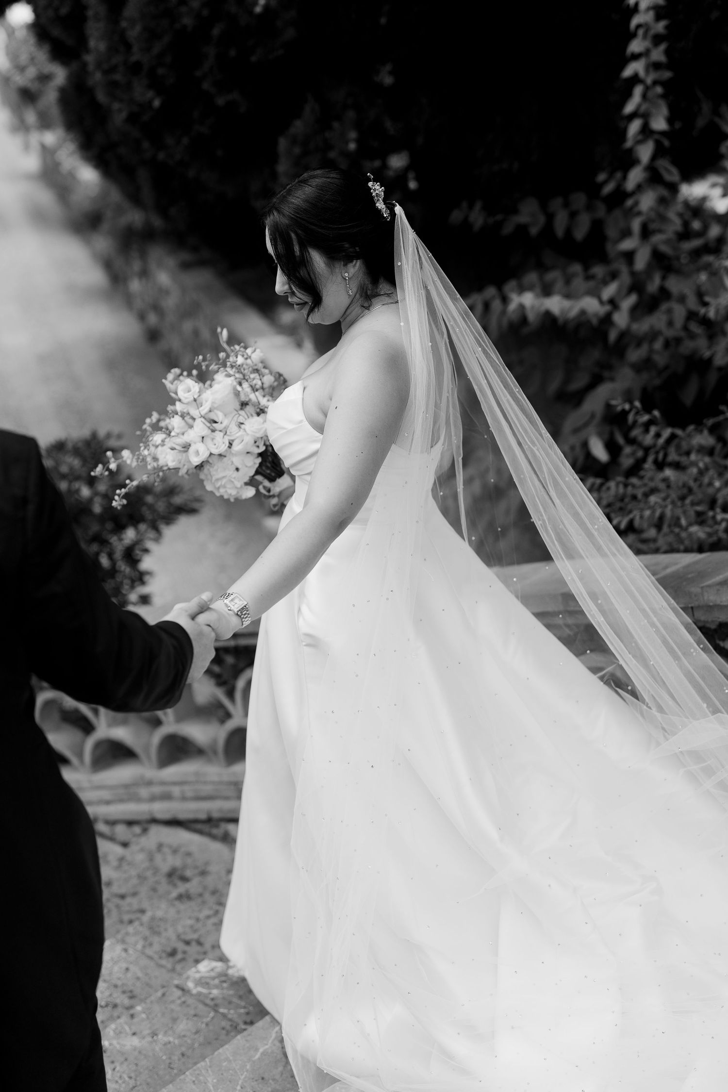 A bride in a flowing white wedding dress and veil walks gracefully through an outdoor garden setting.