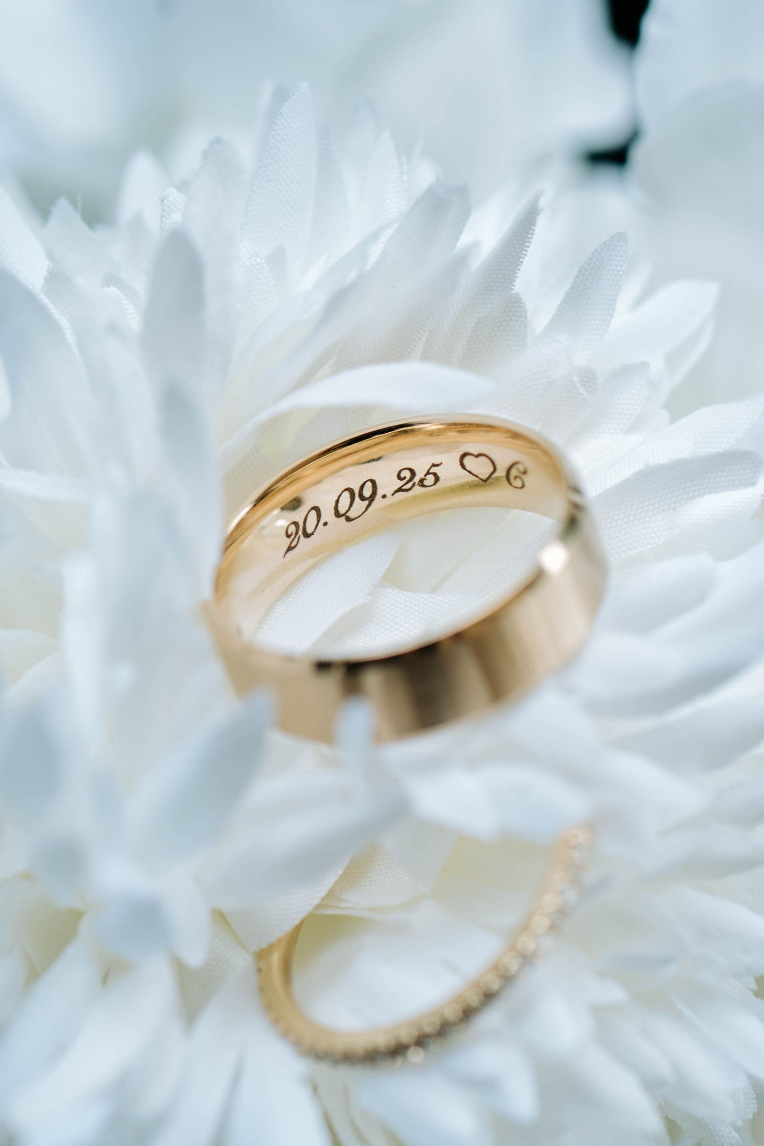Gold wedding ring with engraved date nestled among delicate white flower petals in a soft macro shot.