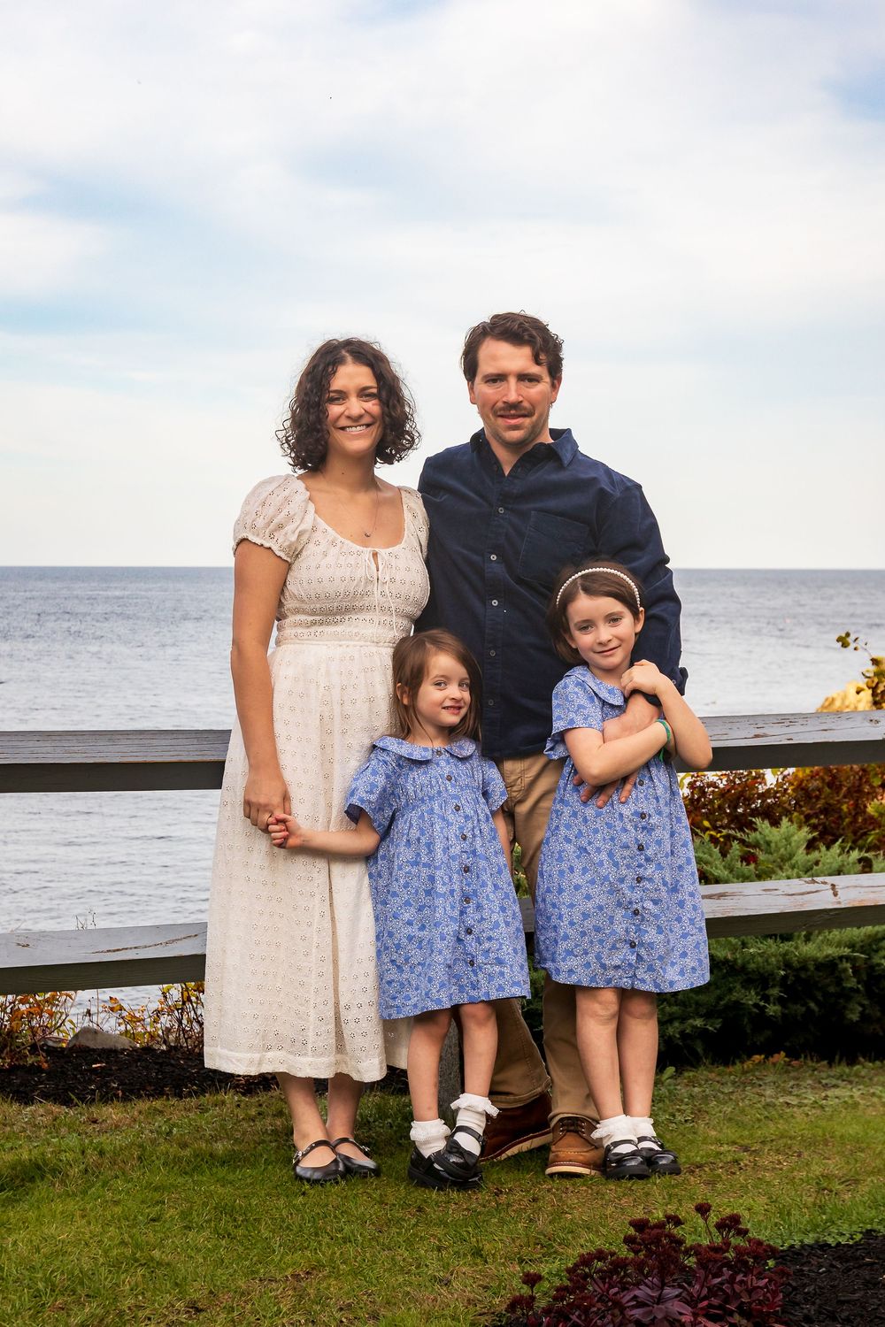 Family in coordinated outfits poses together by the ocean with a white dress and blue floral dresses.