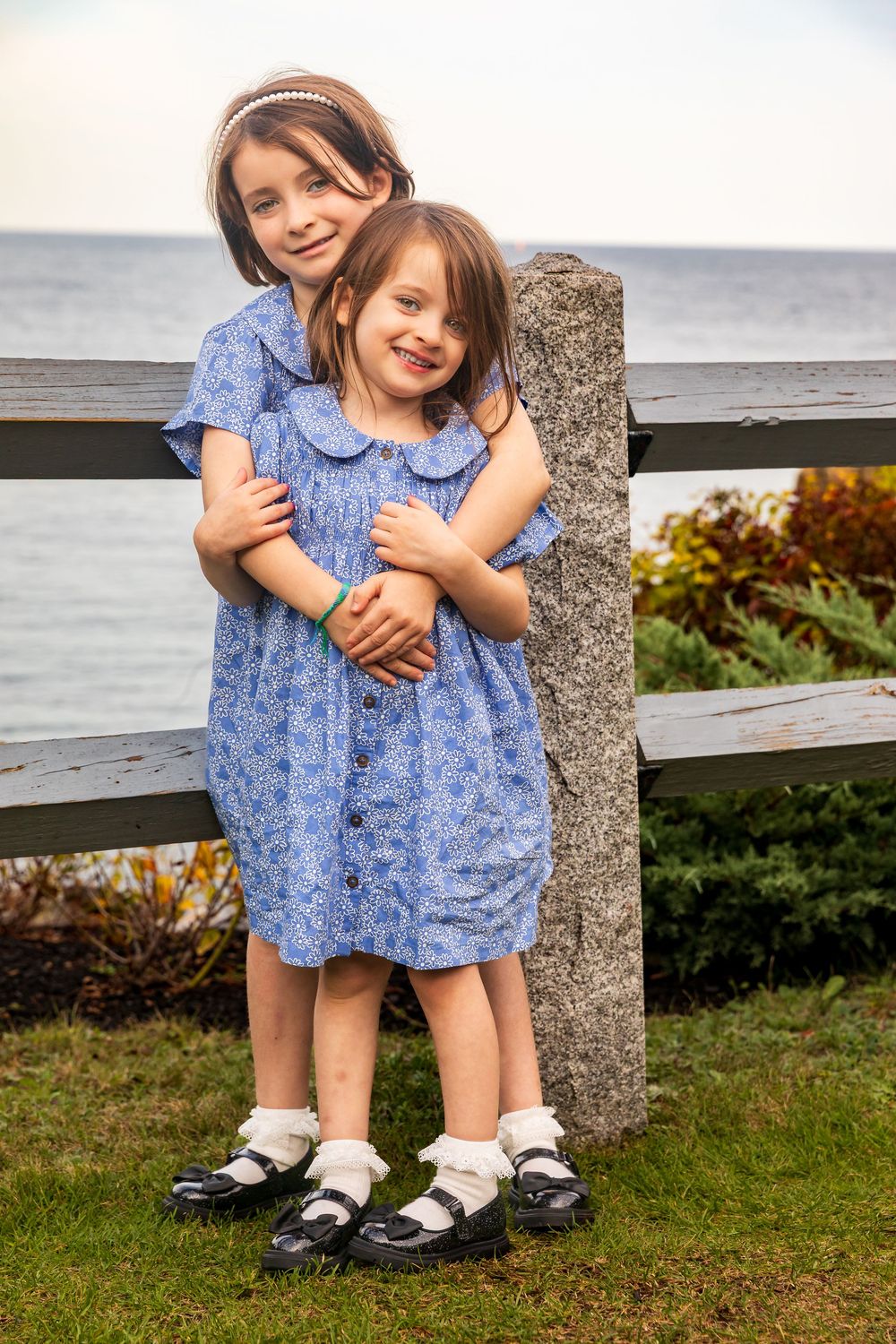 Two children in matching blue floral dresses sit together on coastal rocks.
