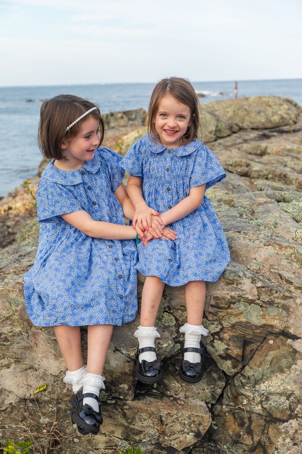 Two children in matching blue dresses sit together on rocky coastal outcrop.