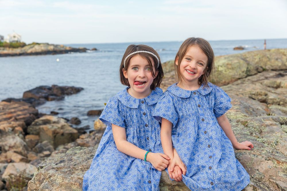 Two children in matching blue floral dresses playing together on rocky coastal shoreline.