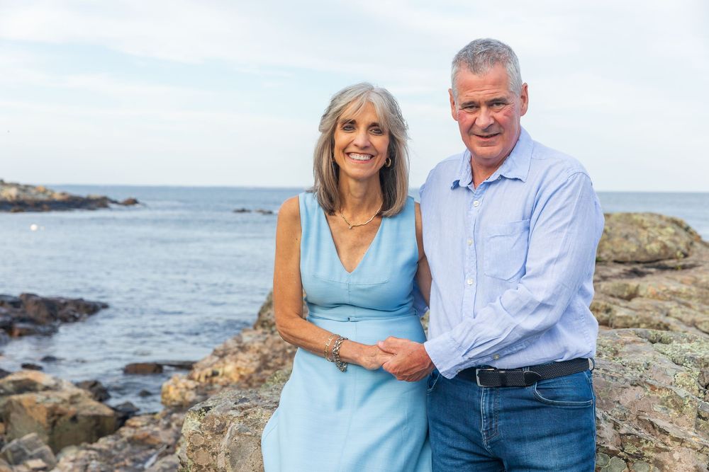 Couple in coordinating blue outfits embrace while standing on rocky ocean shoreline.