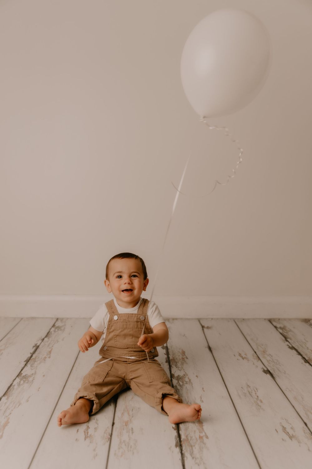 Baby in brown overalls sits on white wooden floor while white balloons float above in minimalist photo series.