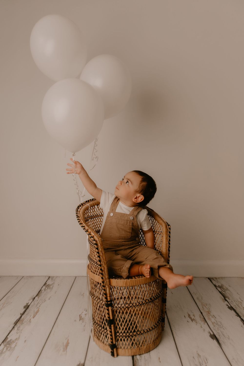Baby in brown wicker chair reaches up toward white balloons against neutral backdrop in studio setting.