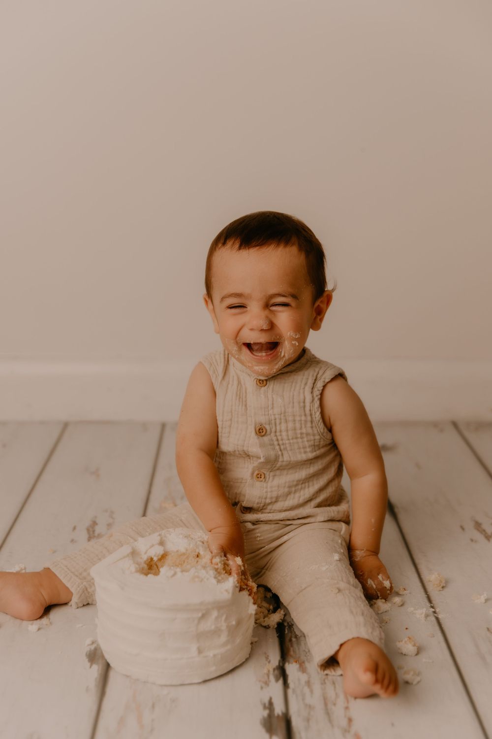 Baby in beige outfit smiles while sitting next to smashed white birthday cake on wooden floor during cake smash photo session.
