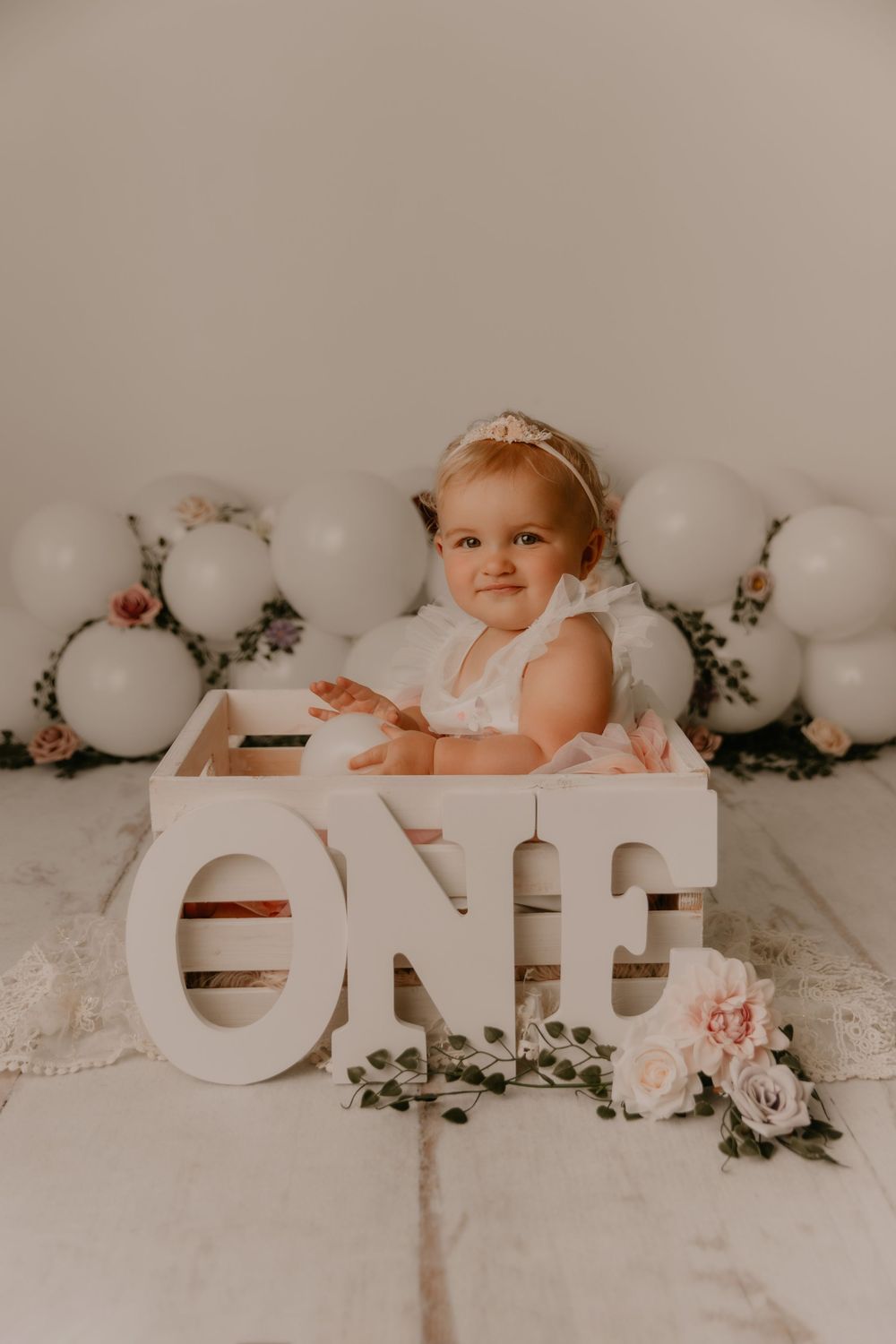 Baby in white dress poses with decorative letters and white balloons against neutral backdrop.