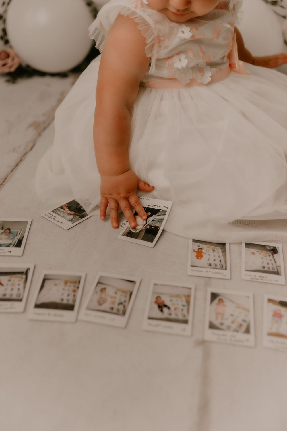 Small hands in a white dress reach down to arrange polaroid photos scattered on a light surface.