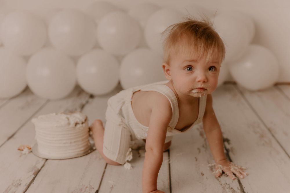 Baby in white outfit sits on floor with cake smash setup featuring white balloons and rustic decorations.