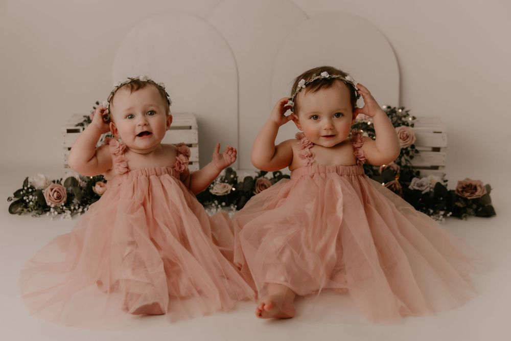 A baby in a pink tulle dress sits and plays on a white floor with floral decorations in the background.