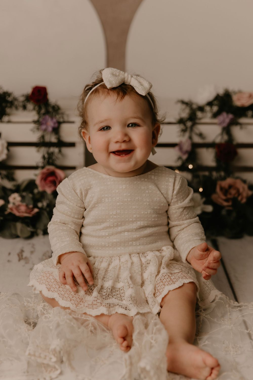 Baby wearing vintage white lace dress sits and smiles in studio setting with floral backdrop.