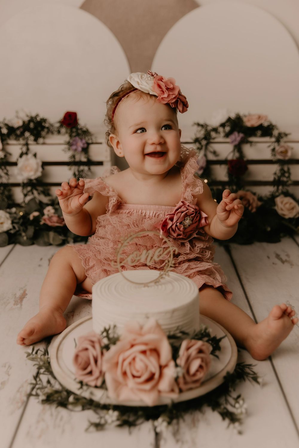 A series of cake smash photos showing a baby in pink ruffled dress celebrating with a white frosted cake and floral decor.