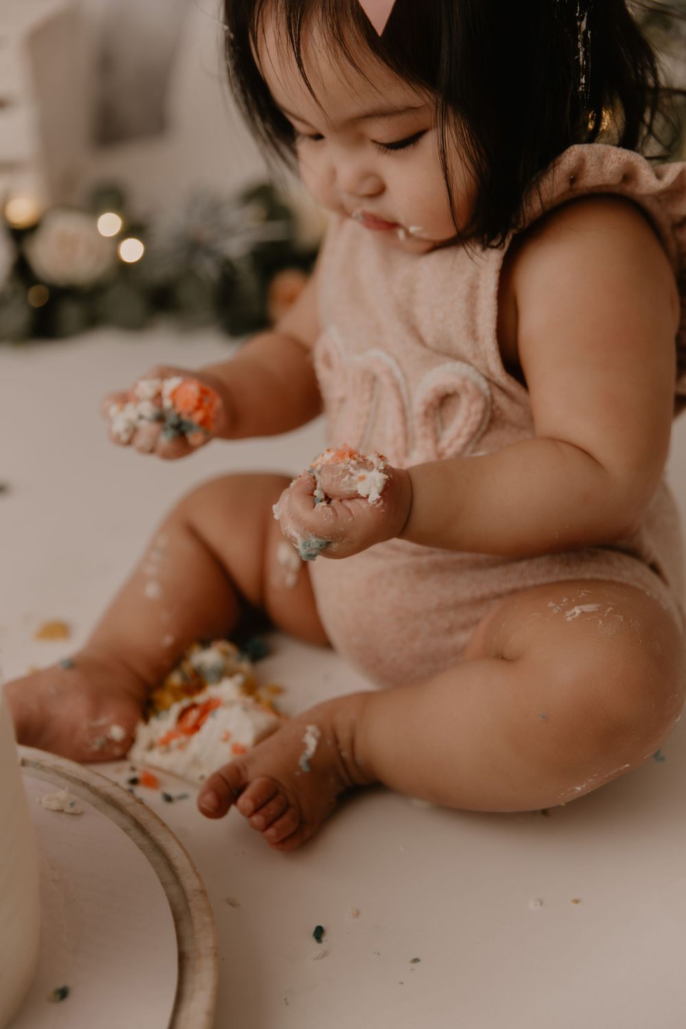 Baby in pink dress sits at table enjoying cake smash photo session with delicate lighting and bokeh background.