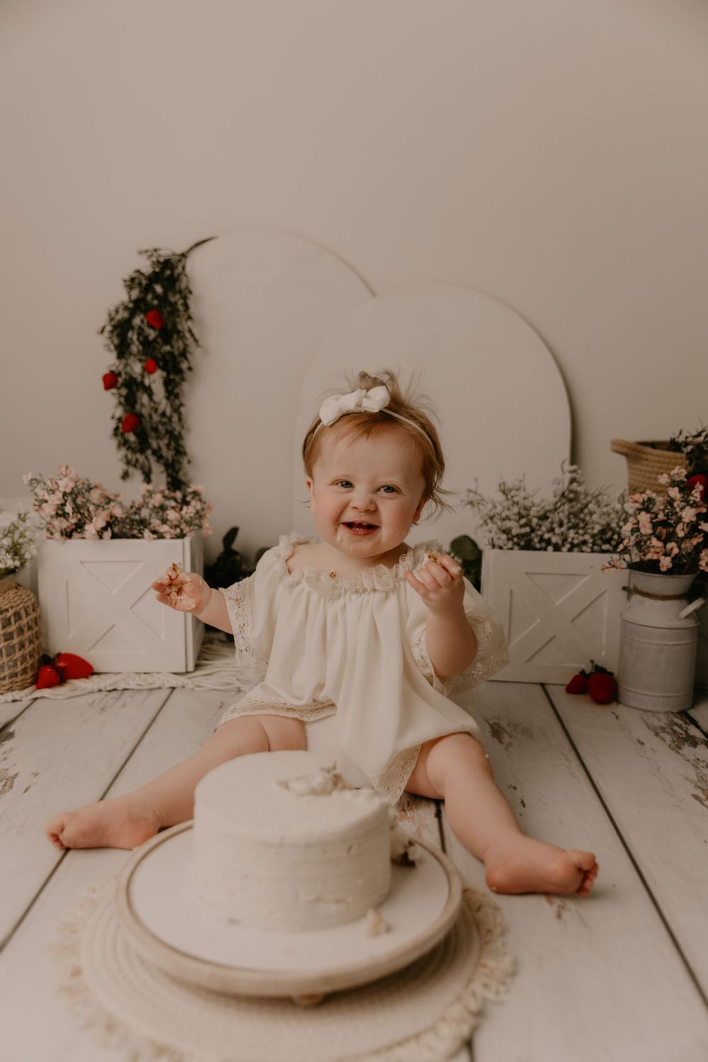 Baby in white dress sits on wooden floor with cake smash setup featuring floral decorations and rustic decor.