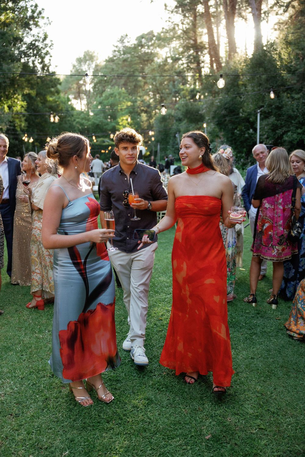 Wedding guests mingle and dance on a grassy lawn at an outdoor reception during sunset.