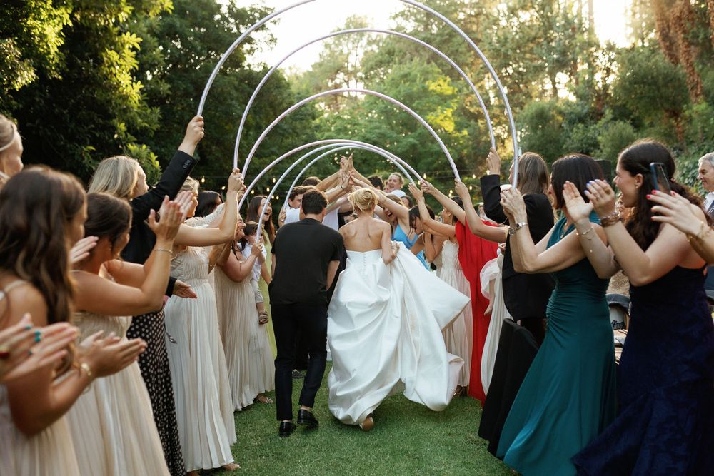 Wedding party members stand in a line outdoors creating an archway with raised arms at sunset.
