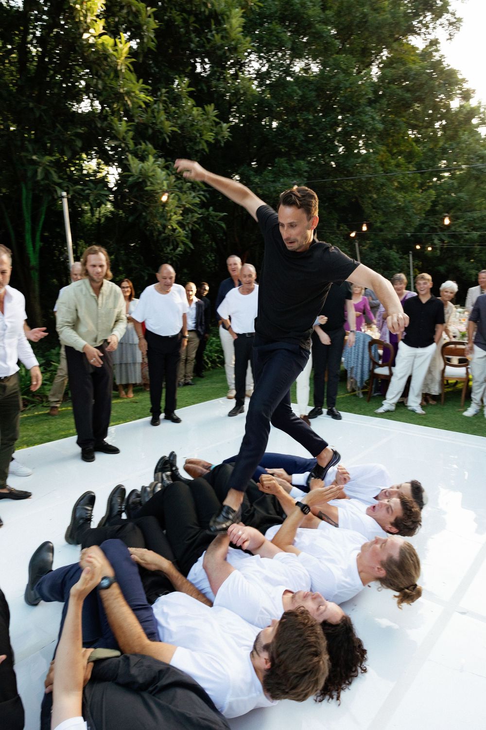 Wedding guests perform a synchronized dance move lying on a white dance floor at an outdoor reception.