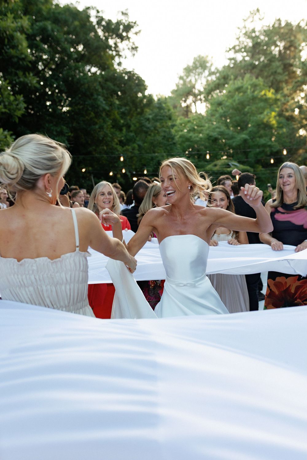 Wedding guests in white dresses create a flowing wave effect during an outdoor celebration.