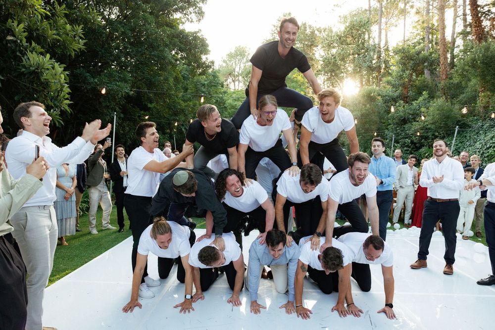 Group of wedding guests perform a synchronized dance move in white attire outdoors.