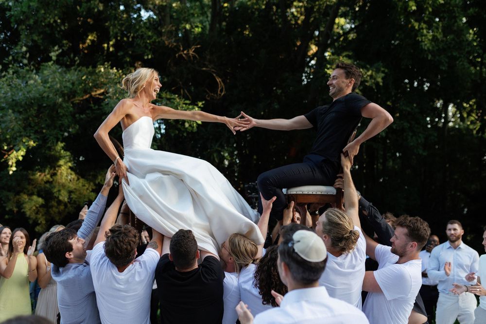 Wedding guests lift a bride and groom in the air during a joyous outdoor celebration.