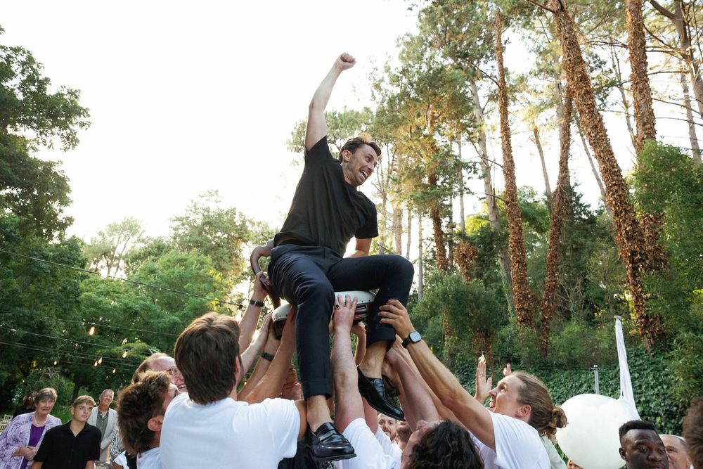 Wedding party dancing and celebrating under tall pine trees at an outdoor reception.