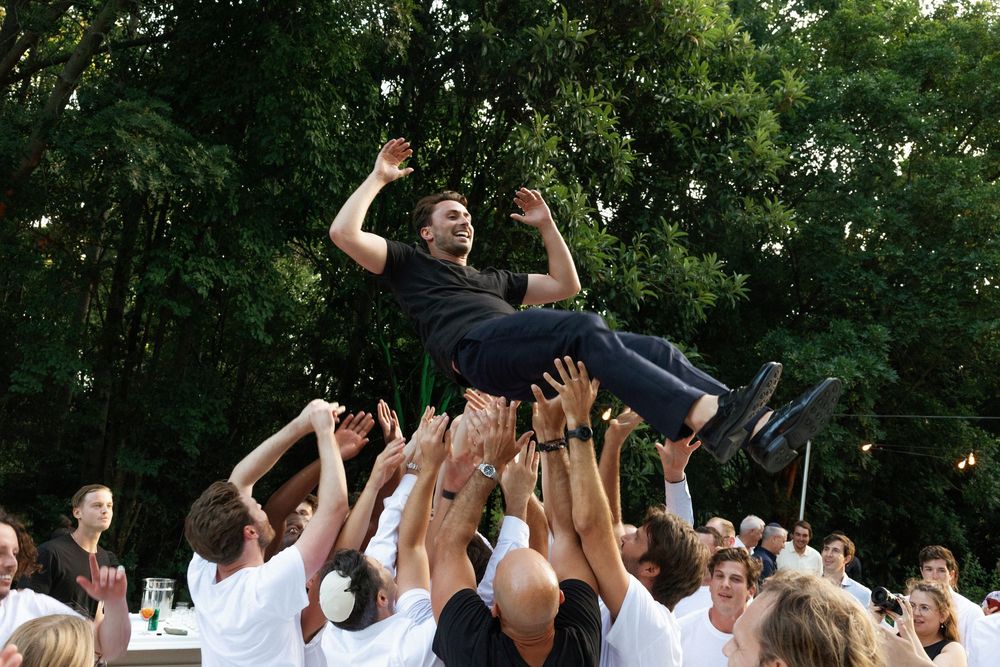 Large group celebrating outdoors with hands raised in the air under green trees.