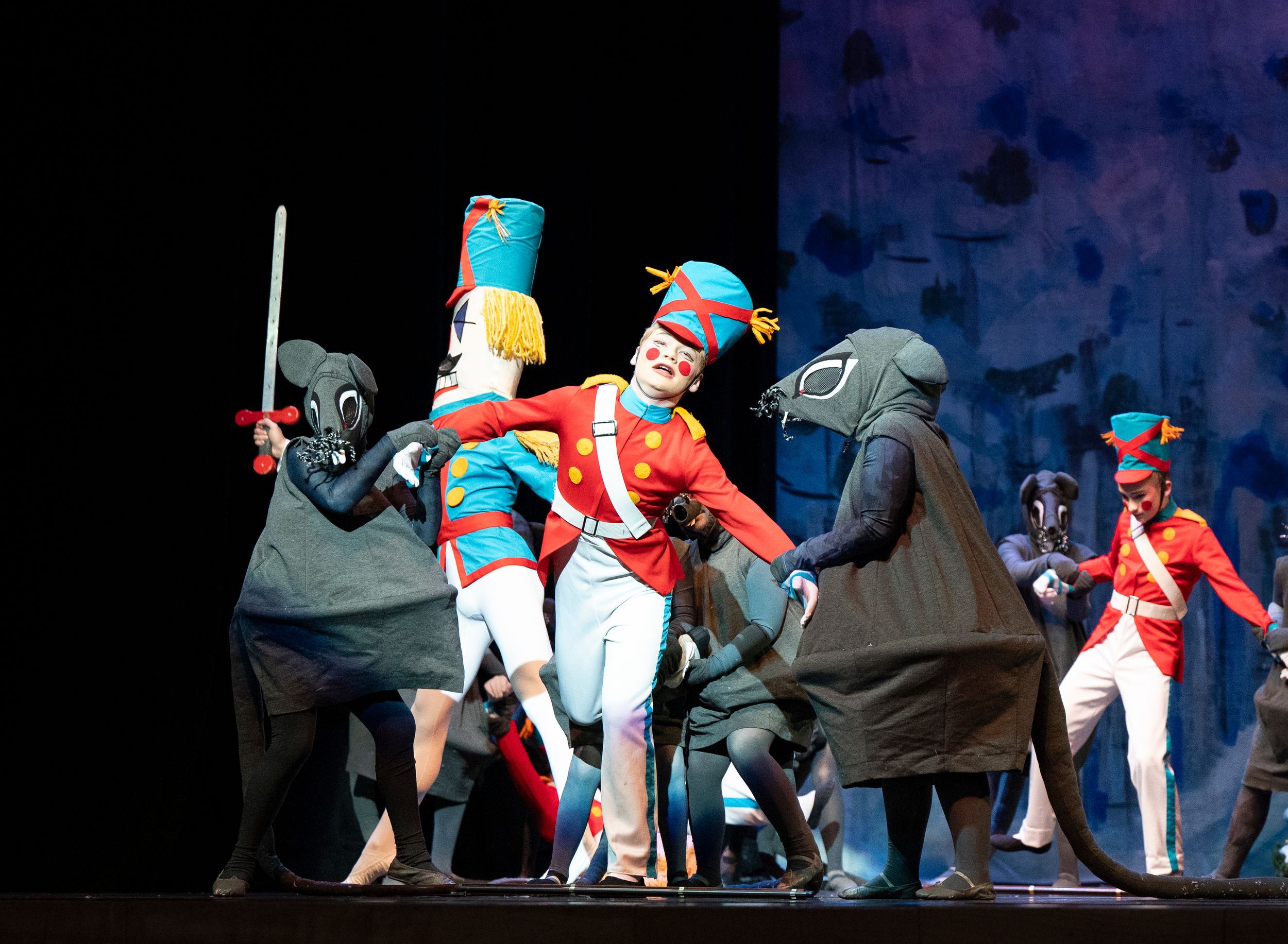 Performers in colorful costumes dance and move across a dark stage during a theatrical production.